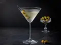 Ein klassischer martini trocken, garniert mit Oliven, steht auf einem dunklen Tisch. Im Hintergrund eine Schale mit weiteren Oliven.