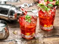 Zwei erfrischende Cocktails mit Erdbeeren und Minze, bereit zum Genießen. Dazu ein Shaker und Eiswürfel auf Holztisch. Perfekt für gute Cocktails.