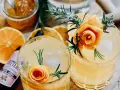 Zwei erfrischende Cocktails mit Orangenscheiben, Rosmarin und einer Orangenschalenrose. Daneben eine Flasche "Bitters" mit Lavendelgeschmack.