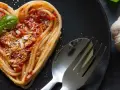 Herzförmige Spaghetti mit Tomatensoße und Basilikum, ein liebevolles Gericht für Pasta Rezepte. Daneben liegen Knoblauch und Tomaten.