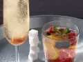 Sektglas mit sprudelndem Champagner-Cocktail, daneben ein Glas mit Beeren, Zitrone und Minze. Ein Stapel Zuckerwürfel.