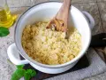 Frisch zubereiteter Couscous in einem Topf, bereit zum Servieren. Ein Holzlöffel lockert die Körner auf. Daneben eine Flasche Öl und Minzblätter.