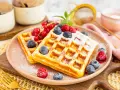 Zwei goldbraune Proteinwaffeln, bestäubt mit Puderzucker und garniert mit frischen Beeren und Johannisbeeren.