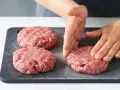 Hände formen rohe Burger-Patties auf einem dunklen Schneidebrett.
