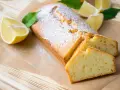 Saftiger veganer Zitronenkuchen, bestäubt mit Puderzucker, neben frischen Zitronenspalten und grünen Blättern.