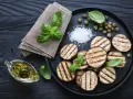 Gegrillte Auberginenscheiben mit Basilikum, Kapern und grobem Salz auf schwarzem Holz. Perfekt zum Aubergine grillen.