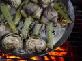 Artischocken werden in einer Pfanne über offenem Feuer zubereitet, brutzeln in Flüssigkeit und duften herrlich.