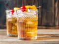 Zwei klassische Gin Old Fashioned Cocktails, garniert mit Kirschen und Orangenzesten, auf einem Holztisch.