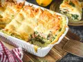 Herzhafte Lachs Spinat Lasagne, goldbraun überbacken, dampft in einer weißen Auflaufform. Ein Stück auf einem Teller zeigt die Schichten.