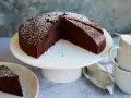 Ein saftiger veganer Schokokuchen, bestäubt mit Puderzucker, wartet auf Genuss. Daneben liegen zwei Tassen.