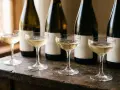 Vier Gläser mit sprudelndem Champagner Sekt stehen auf einem Holztisch vor mehreren dunklen Flaschen.
