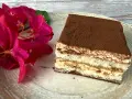 Ein Stück Tiramisu, perfekt für Ihr nächstes tiramisu rezept, liegt auf einem Teller neben leuchtend roten Blüten.