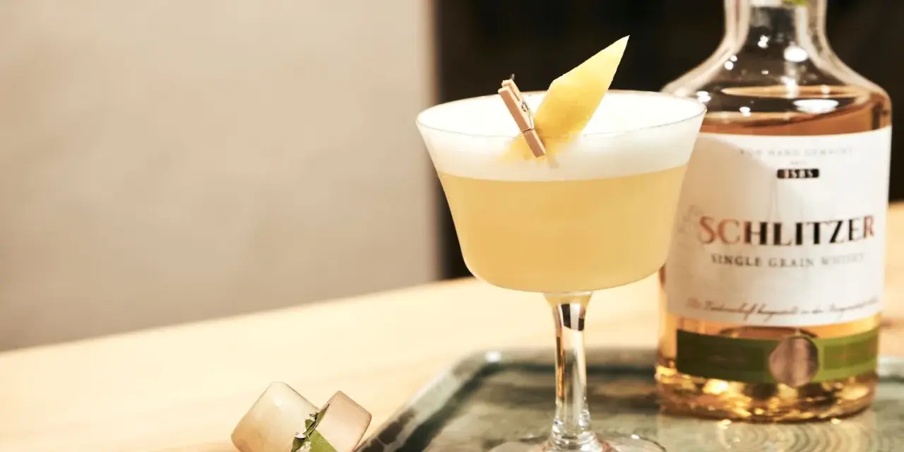 Ein erfrischender Sour Cocktail mit Zitronenspalte, neben einer Flasche Schlitzer Single Grain Whisky. Perfekt für ein Sour Cocktail Rezept.