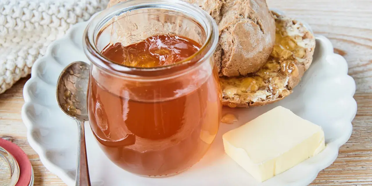 Glas mit goldbraunem Apfelgelee aus Apfelsaft, daneben ein Stück Butter und ein halbes Brot auf einem Teller.