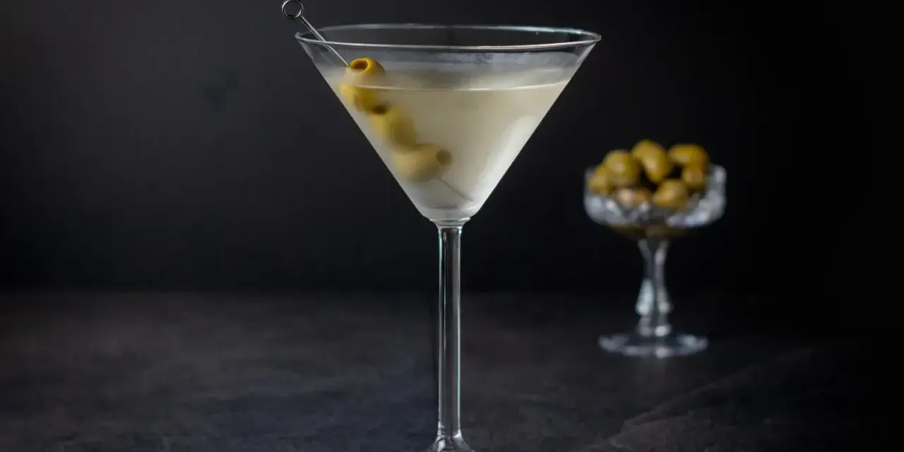 Ein klassischer martini trocken, garniert mit Oliven, steht auf einem dunklen Tisch. Im Hintergrund eine Schale mit weiteren Oliven.