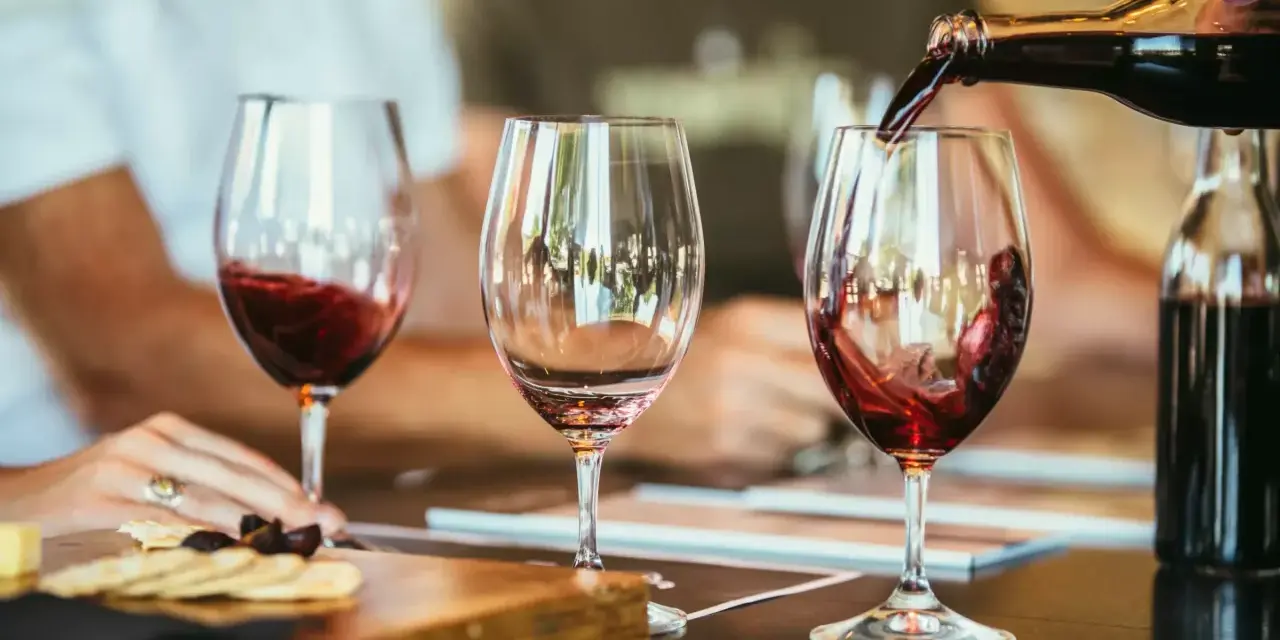 Rotwein wird in ein Glas gegossen, der Duft von **Wein** und **Tanninen** liegt in der Luft. Daneben ein leeres Glas und ein Teller mit Käse und Crackern.