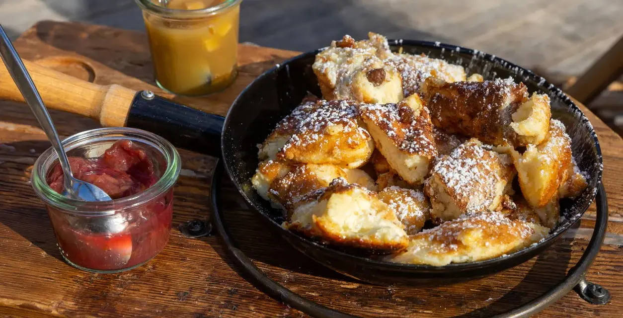 Ein frisch zubereiteter Kaiserschmarrn im Pfännchen, bestäubt mit Puderzucker, daneben Apfelmus und Zwetschgenröster. Ein leckeres Kaiserschmarrn Rezept zum Nachmachen.