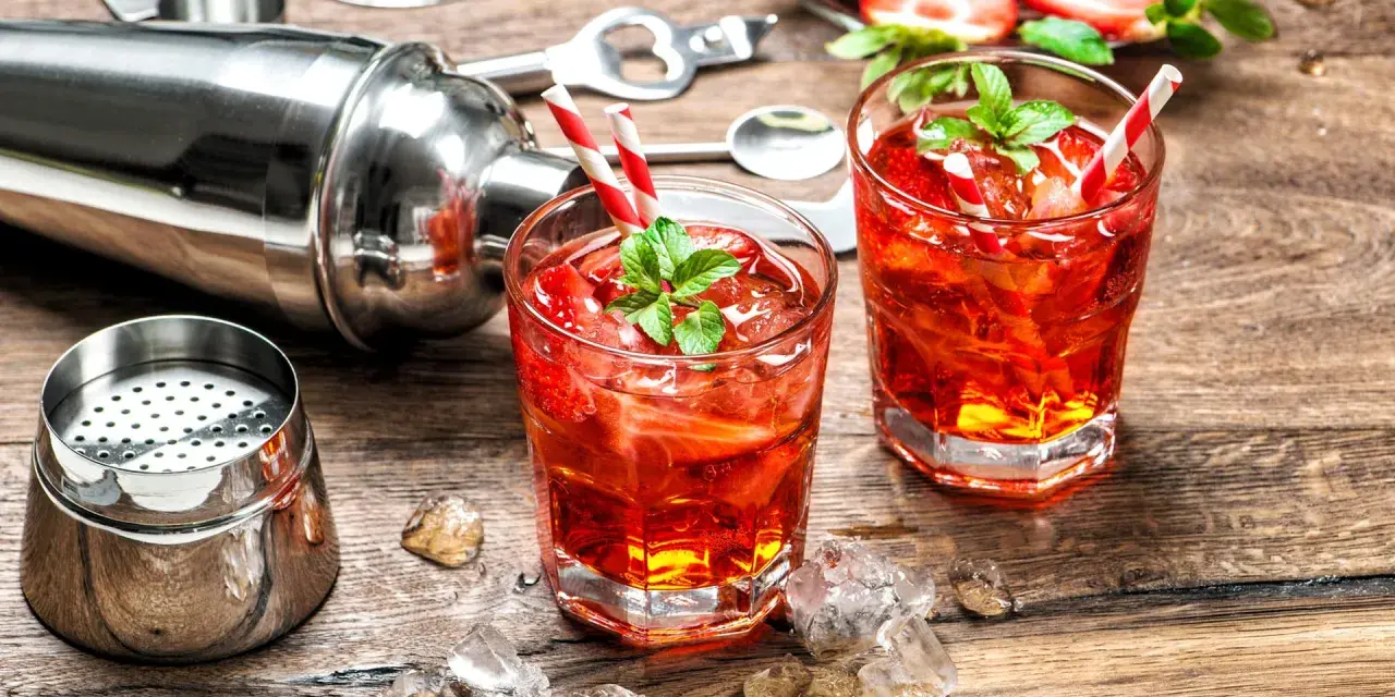 Zwei erfrischende Cocktails mit Erdbeeren und Minze, bereit zum Genießen. Dazu ein Shaker und Eiswürfel auf Holztisch. Perfekt für gute Cocktails.
