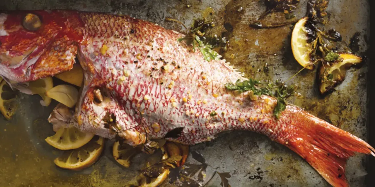 Ein ganzer, gebackener Red Snapper mit Zitronenspalten und Kräutern auf einem Backblech.