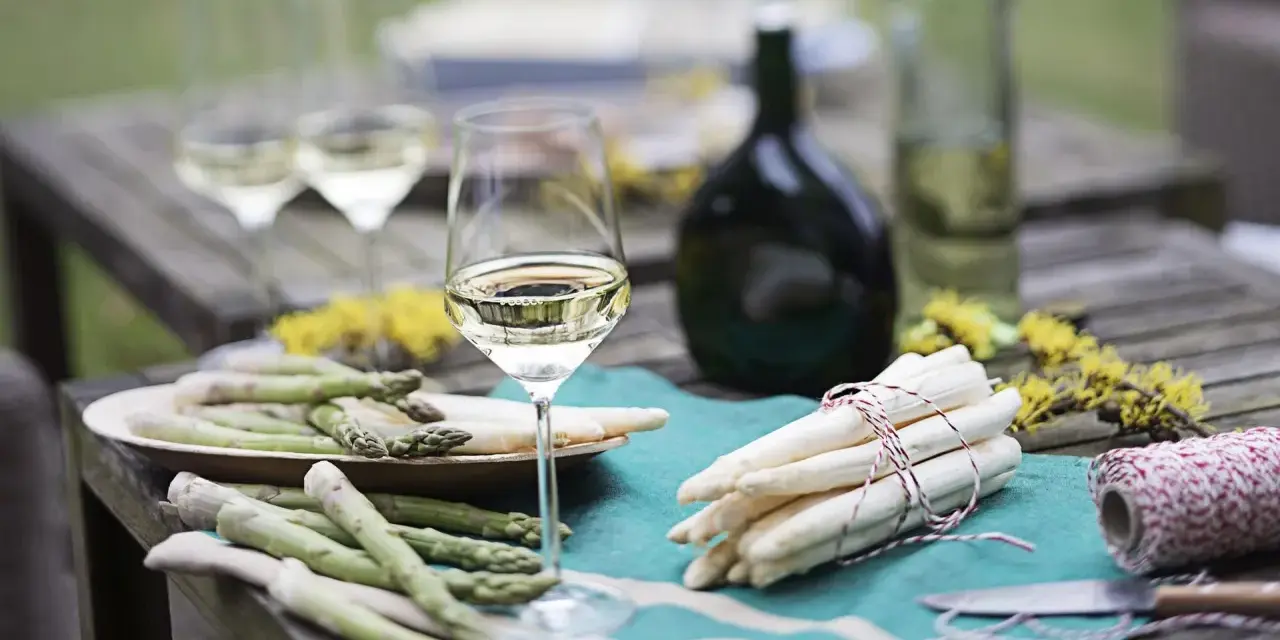 Ein Glas Weißwein steht neben frischem Spargel auf einem Holztisch. Die Frage welcher Wein zum Spargel passt, wird hier visuell beantwortet.