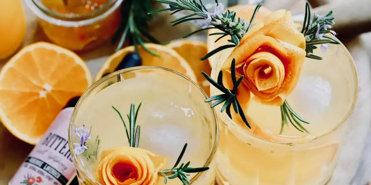 Zwei erfrischende Cocktails mit Orangenscheiben, Rosmarin und einer Orangenschalenrose. Daneben eine Flasche "Bitters" mit Lavendelgeschmack.