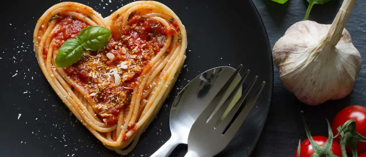 Herzförmige Spaghetti mit Tomatensoße und Basilikum, ein liebevolles Gericht für Pasta Rezepte. Daneben liegen Knoblauch und Tomaten.