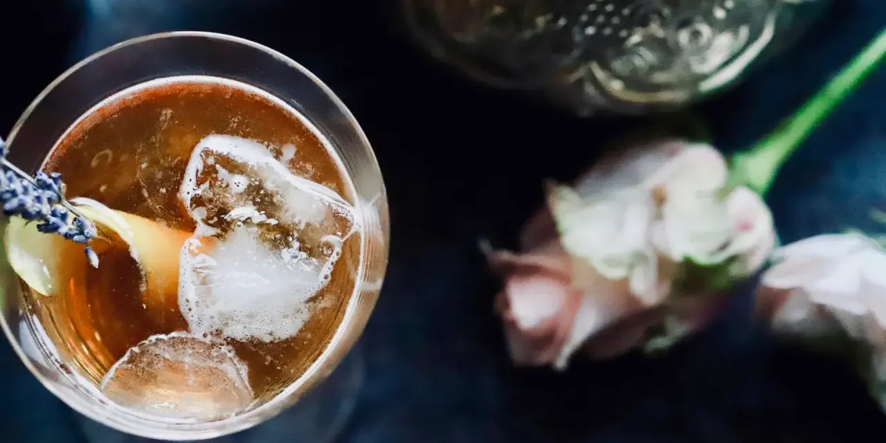 Ein erfrischender Gin Tee mit Eis, Zitrone und Lavendelblüten. Daneben zarte Rosen und eine verzierte Karaffe.