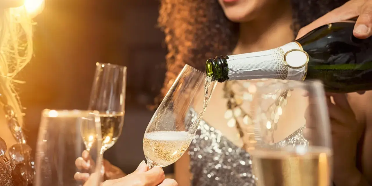 Sekt wird in ein Glas gegossen, während Freunde anstoßen. Der Unterschied zwischen Wein und Sekt ist die Kohlensäure, die Sekt prickelnd macht.