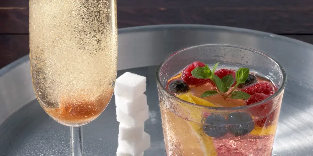 Sektglas mit sprudelndem Champagner-Cocktail, daneben ein Glas mit Beeren, Zitrone und Minze. Ein Stapel Zuckerwürfel.