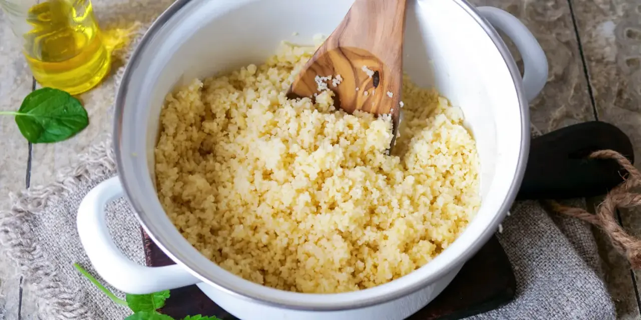 Frisch zubereiteter Couscous in einem Topf, bereit zum Servieren. Ein Holzlöffel lockert die Körner auf. Daneben eine Flasche Öl und Minzblätter.