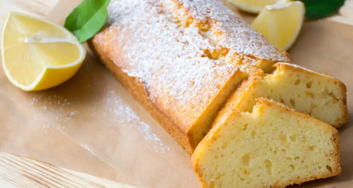 Saftiger veganer Zitronenkuchen, bestäubt mit Puderzucker, neben frischen Zitronenspalten und grünen Blättern.