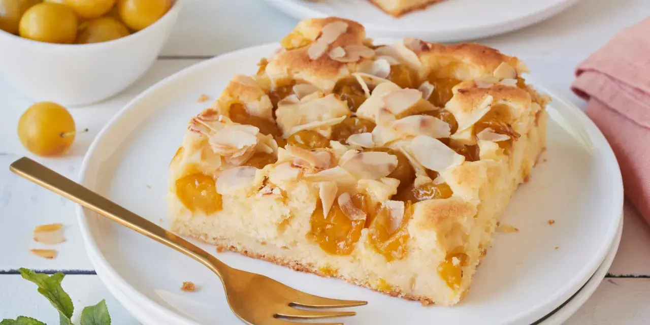 Ein Stück saftiger Mirabellenkuchen mit Mandelblättchen, serviert auf einem Teller mit einer goldenen Gabel. Daneben eine Schale voller frischer Mirabellen.