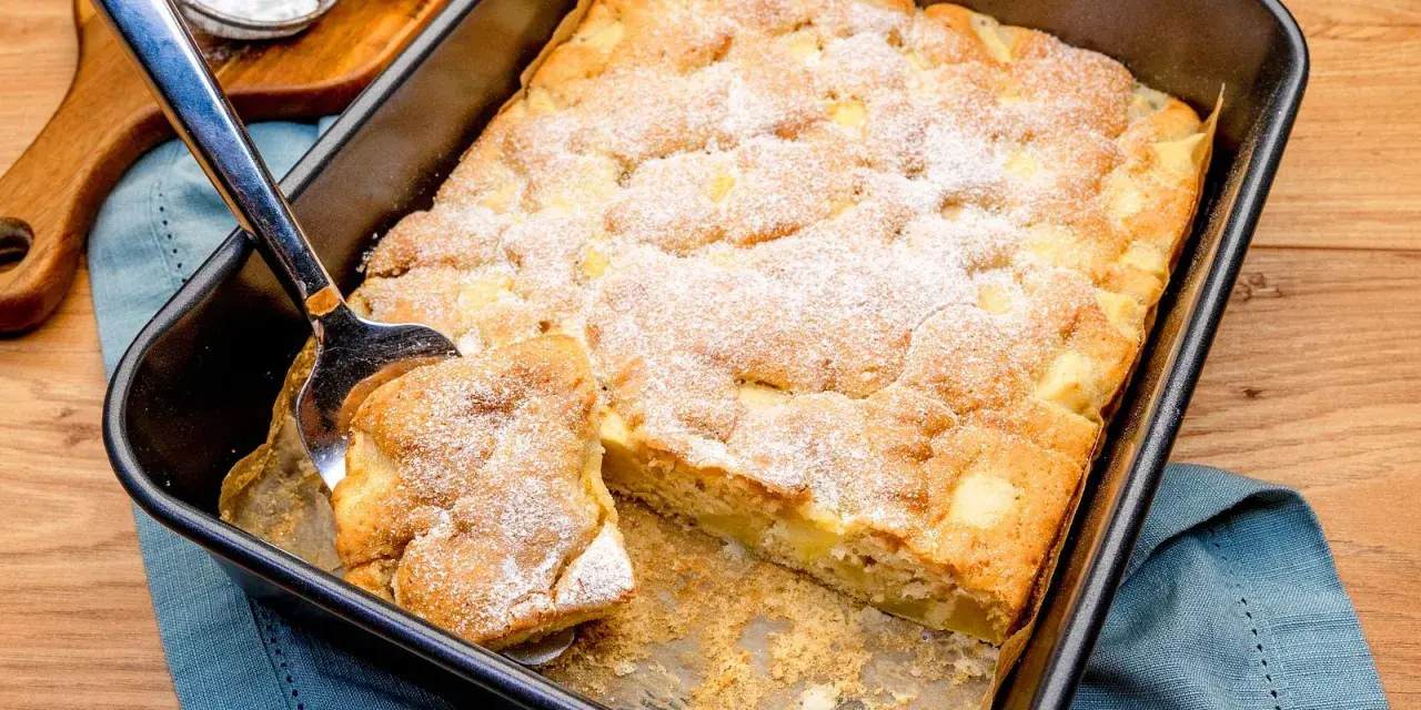 Ein Stück vom schneller saftiger apfelkuchen vom blech wird mit einem Kuchenheber herausgehoben. Mit Puderzucker bestäubt.