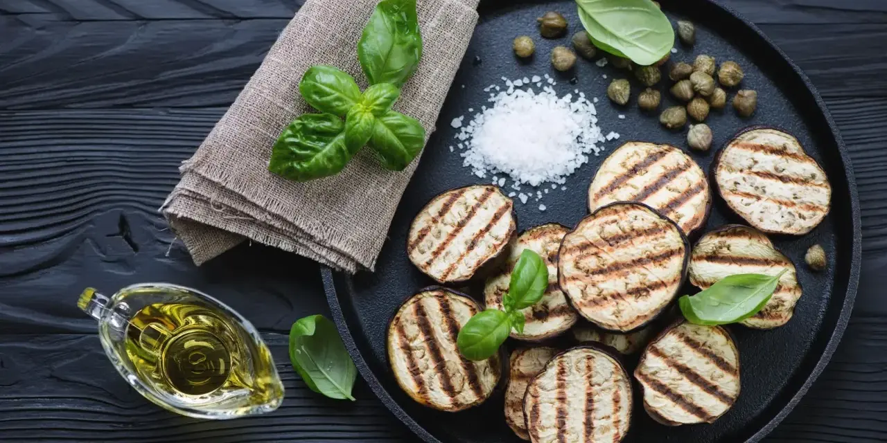 Gegrillte Auberginenscheiben mit Basilikum, Kapern und grobem Salz auf schwarzem Holz. Perfekt zum Aubergine grillen.