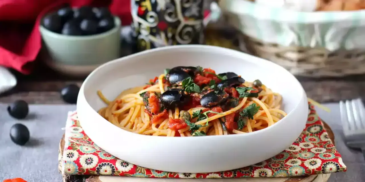 Eine Schüssel Spaghetti Puttanesca, garniert mit schwarzen Oliven und Kräutern, auf einem rustikalen Holzständer.