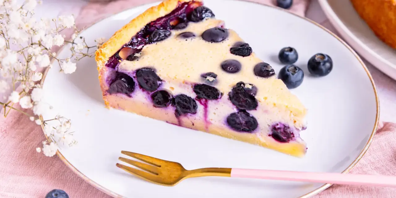 Ein Stück saftiger Heidelbeerkuchen Landfrauenart auf einem Teller, garniert mit frischen Beeren und einer Gabel.