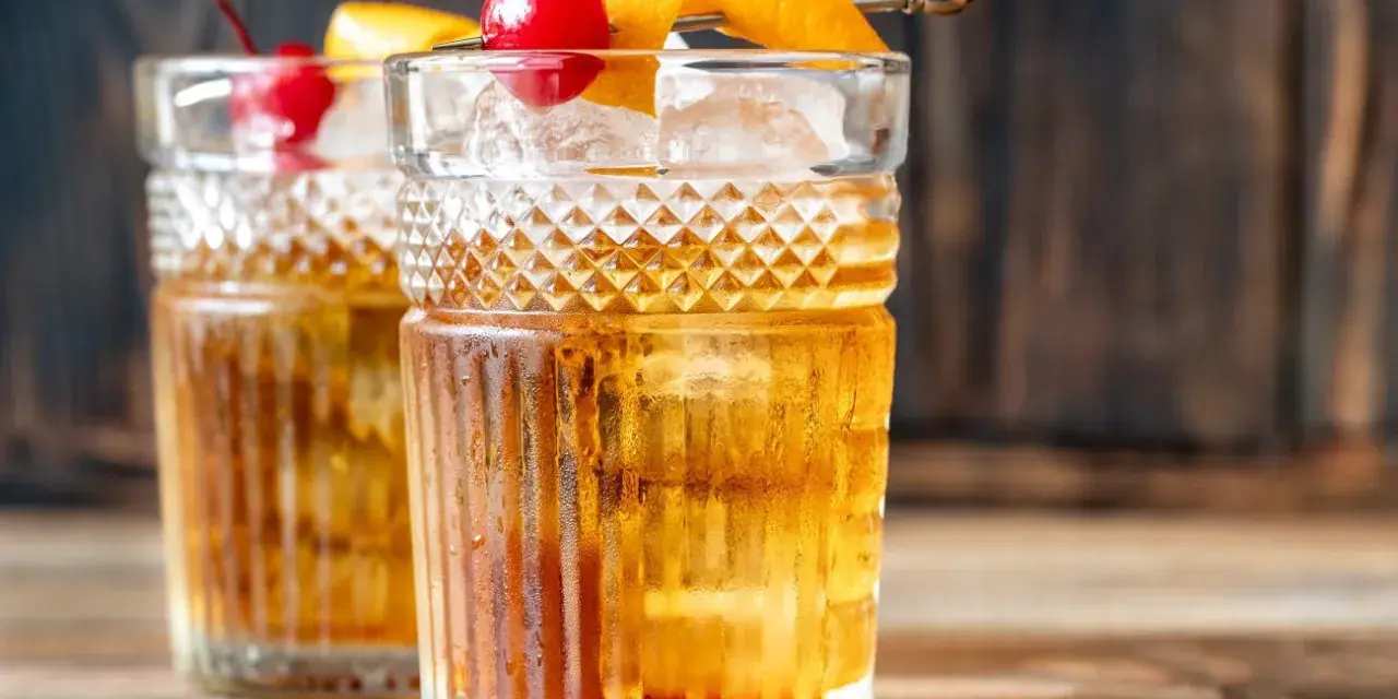 Zwei klassische Gin Old Fashioned Cocktails, garniert mit Kirschen und Orangenzesten, auf einem Holztisch.