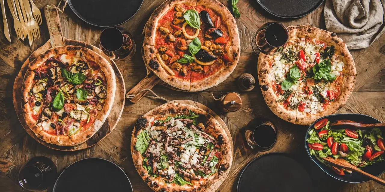 Vier leckere Pizzen und ein Salat auf Holztisch. Die Frage welcher Wein zu Pizza passt, wird hier mit Rotwein beantwortet.