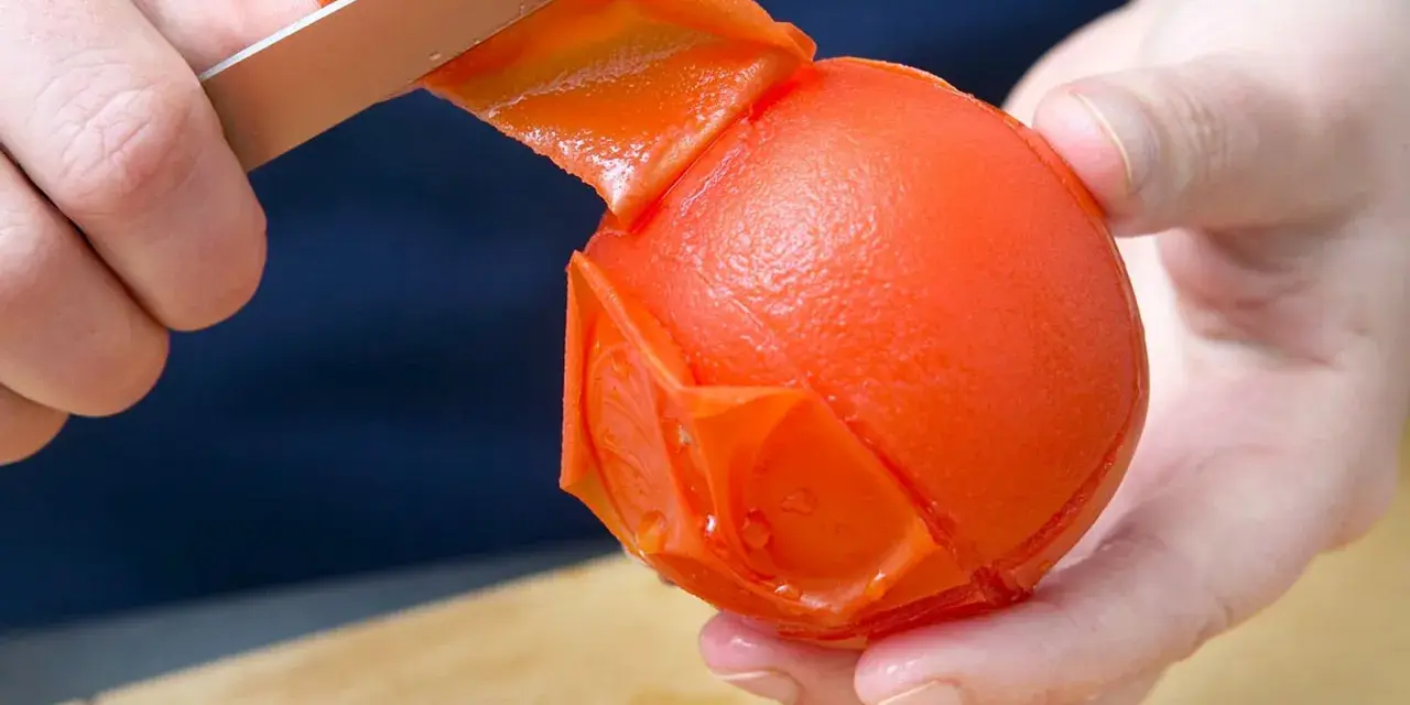 Hände ziehen mit einem Messer die Haut von einer Tomate ab. Die Tomate ist fast komplett geschält.