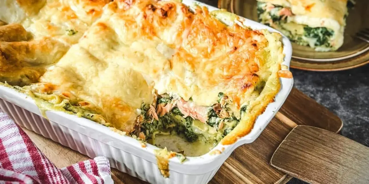 Herzhafte Lachs Spinat Lasagne, goldbraun überbacken, dampft in einer weißen Auflaufform. Ein Stück auf einem Teller zeigt die Schichten.