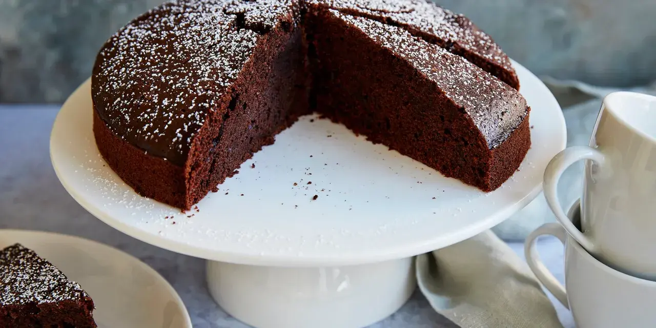 Ein saftiger veganer Schokokuchen, bestäubt mit Puderzucker, wartet auf Genuss. Daneben liegen zwei Tassen.