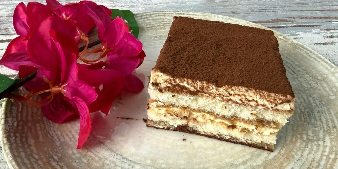 Ein Stück Tiramisu, perfekt für Ihr nächstes tiramisu rezept, liegt auf einem Teller neben leuchtend roten Blüten.