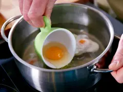 Ein rohes Ei wird aus einer grünen Tasse in kochendes Wasser gegossen, um verlorene Eier zuzubereiten.