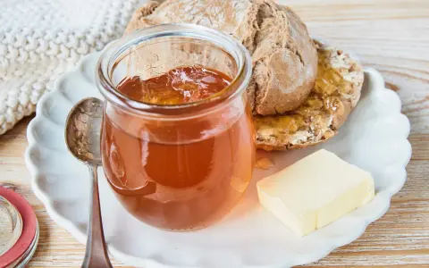 Glas mit goldbraunem Apfelgelee aus Apfelsaft, daneben ein Stück Butter und ein halbes Brot auf einem Teller.