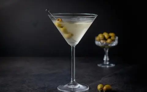 Ein klassischer martini trocken, garniert mit Oliven, steht auf einem dunklen Tisch. Im Hintergrund eine Schale mit weiteren Oliven.