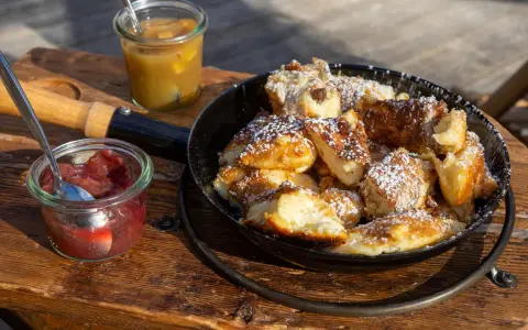 Ein frisch zubereiteter Kaiserschmarrn im Pfännchen, bestäubt mit Puderzucker, daneben Apfelmus und Zwetschgenröster. Ein leckeres Kaiserschmarrn Rezept zum Nachmachen.