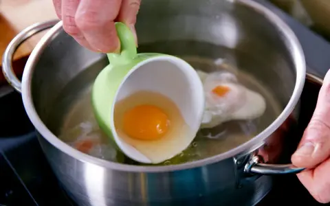 Ein rohes Ei wird aus einer grünen Tasse in kochendes Wasser gegossen, um verlorene Eier zuzubereiten.