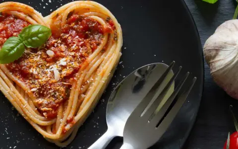 Herzförmige Spaghetti mit Tomatensoße und Basilikum, ein liebevolles Gericht für Pasta Rezepte. Daneben liegen Knoblauch und Tomaten.