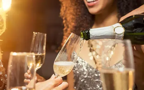 Sekt wird in ein Glas gegossen, während Freunde anstoßen. Der Unterschied zwischen Wein und Sekt ist die Kohlensäure, die Sekt prickelnd macht.