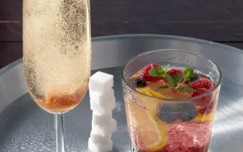 Sektglas mit sprudelndem Champagner-Cocktail, daneben ein Glas mit Beeren, Zitrone und Minze. Ein Stapel Zuckerwürfel.